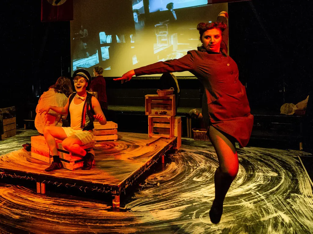 A group of HCA performing arts students mid-performance in the Folly Arts Theatre, two dancers circle around a central raised stage where two actors sit.