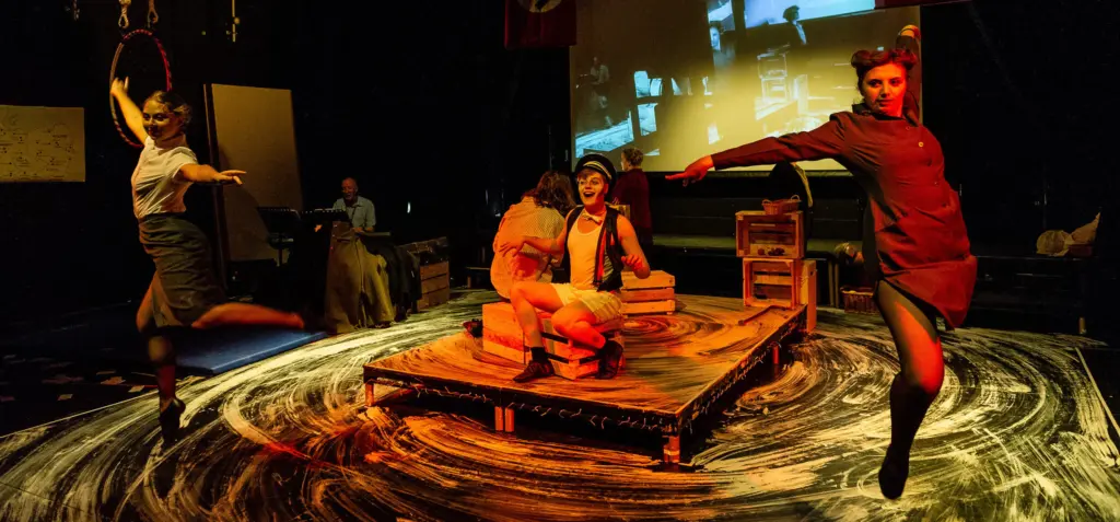 A group of HCA performing arts students mid-performance in the Folly Arts Theatre, two dancers circle around a central raised stage where two actors sit.