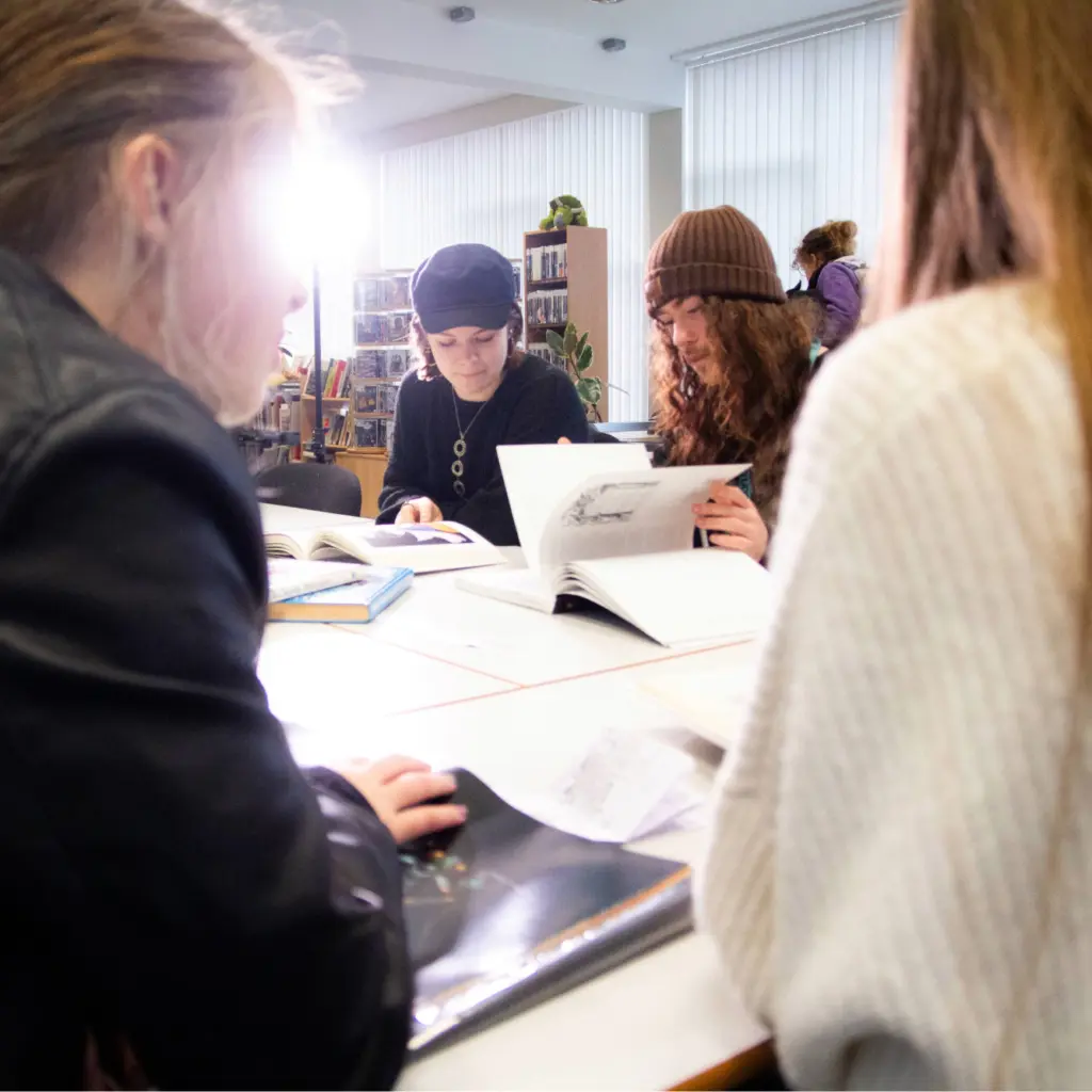 Group of student working in the library around a large square table. Students in the foreground and background using reference books.