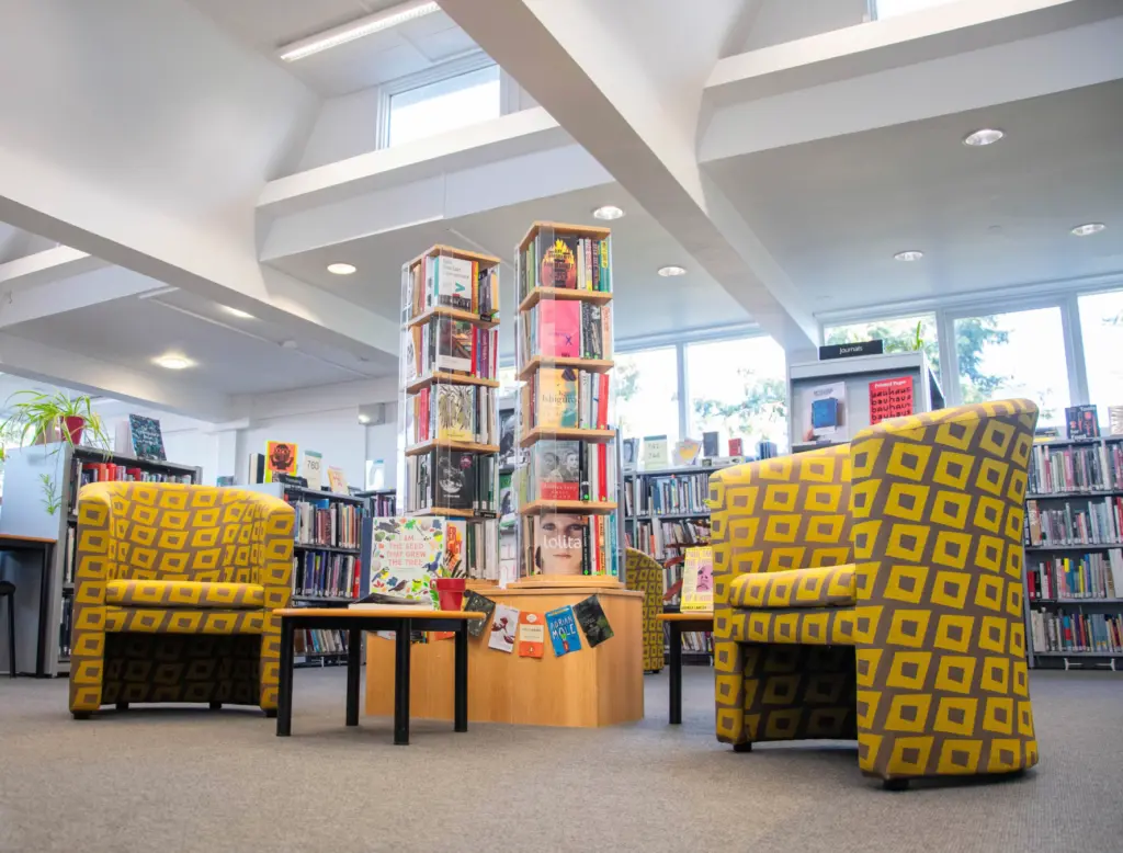 2 bright yellow armchairs in the library. They are spaced apart and are surrounded by colourful books.