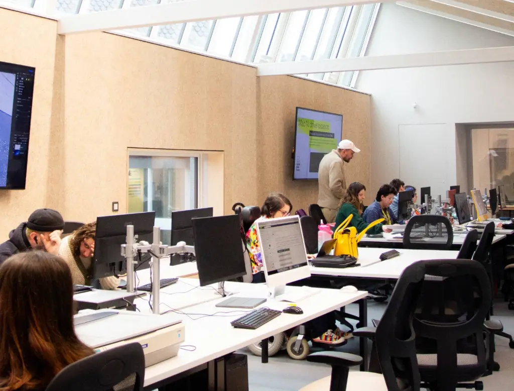 A wide-angle shot of the digital skills centre. A desk with macs and PCs go through the centre of the room, with students at the computers and a tutor standing in front of a screen, looking at a students work.