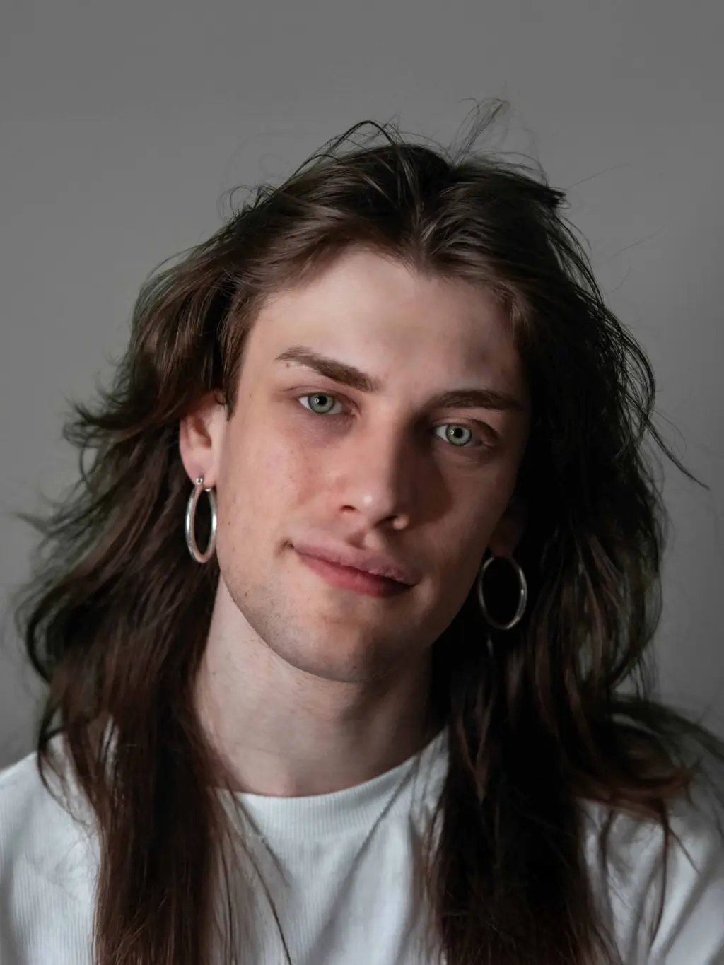 A studio photo of Kit Newman, they are wearing a white t-shirt and hoop earrings and they have long brown hair. They are softly smiling at the camera against a grey background.