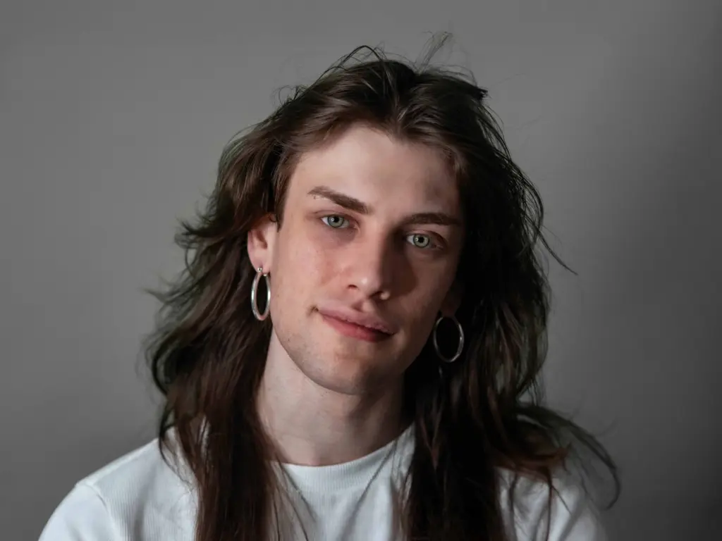 A studio photo of Kit Newman, they are wearing a white t-shirt and hoop earrings and they have long brown hair. They are softly smiling at the camera against a grey background.