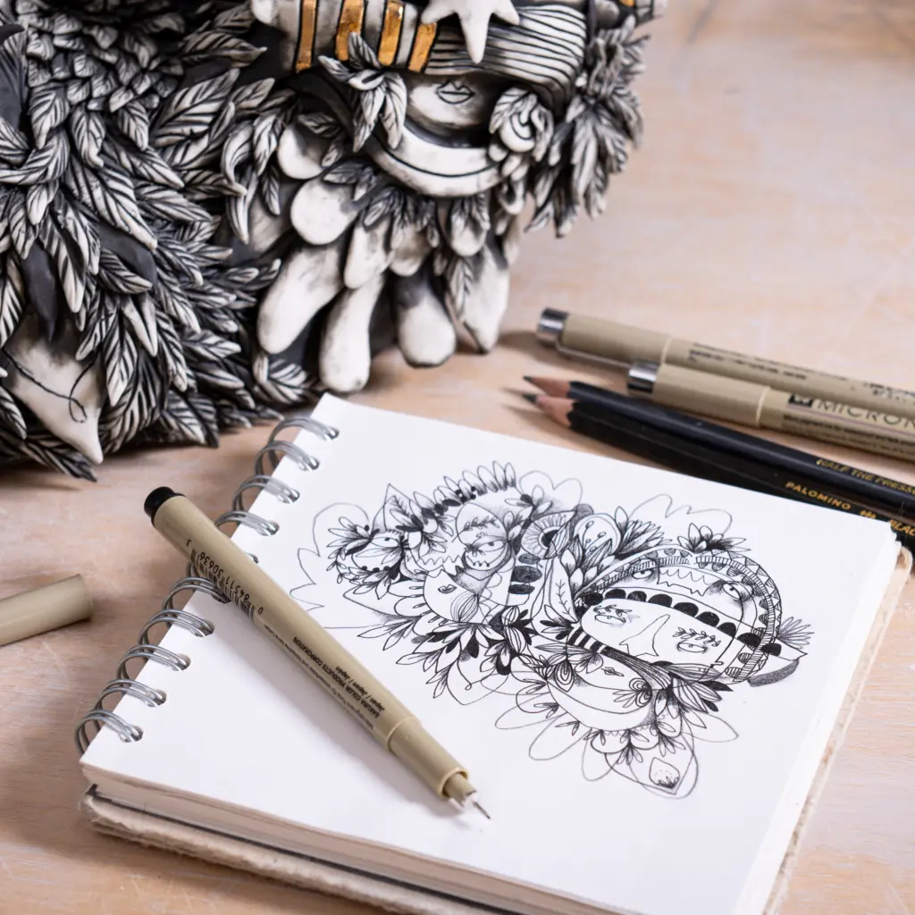 A photo of a sketchbook lying next to a ceramic sculpture. The sketchbook has an illustrative drawing of characters in a unique style and the sculpture is an interpretation of the drawing. There are pens lying on the table next to the sculpture and sketchbook.