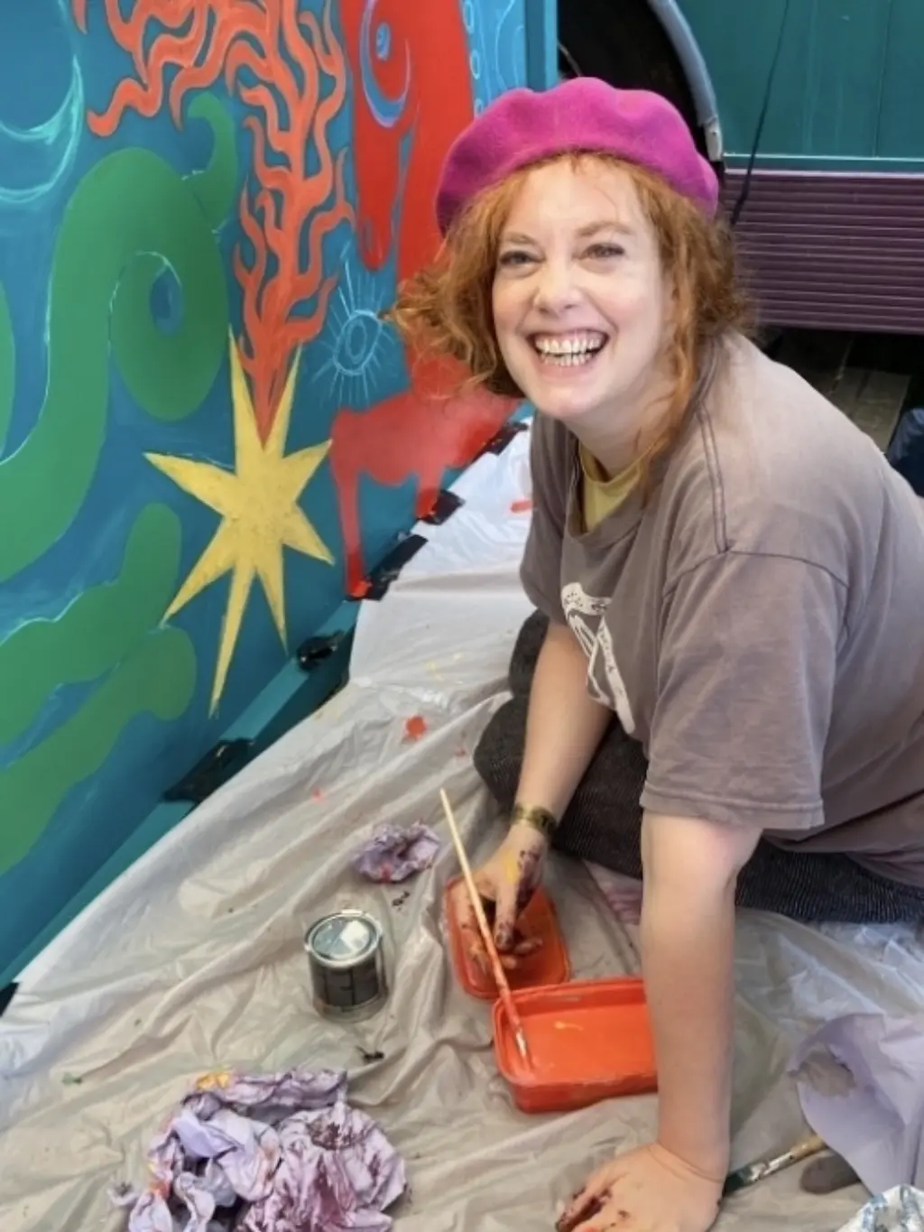 Photo of Lexi Strauss painting a colourful painting. She is wearing a neon pink beret and is smiling at the camera.