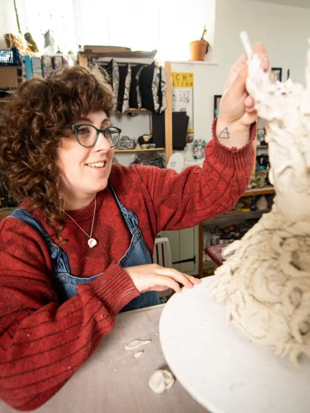 Lucy Baxendale works on a ceramic sculpture in her home studio. She has curly brown hair and glasses, and wears a red jumper with blue dungarees.