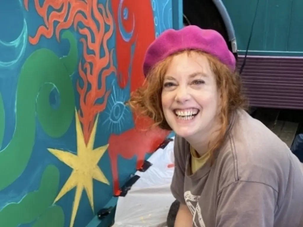 Photo of Lexi Strauss painting a colourful painting. She is wearing a neon pink beret and is smiling at the camera.