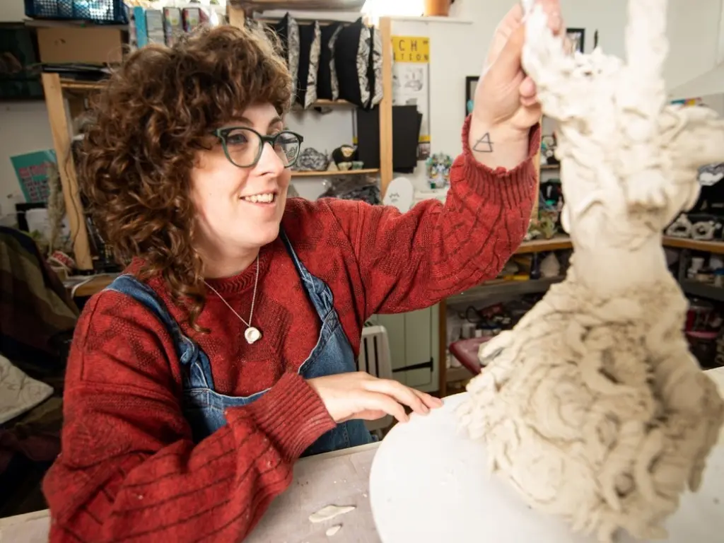 Lucy Baxendale works on a ceramic sculpture in her home studio. She has curly brown hair and glasses, and wears a red jumper with blue dungarees.