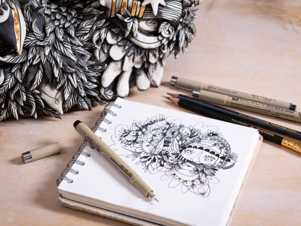 A photo of a sketchbook lying next to a ceramic sculpture. The sketchbook has an illustrative drawing of characters in a unique style and the sculpture is an interpretation of the drawing. There are pens lying on the table next to the sculpture and sketchbook.