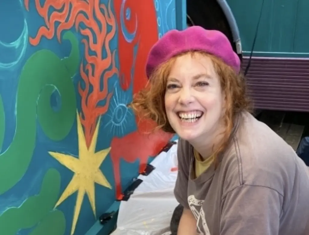 Photo of Lexi Strauss painting a colourful painting. She is wearing a neon pink beret and is smiling at the camera.