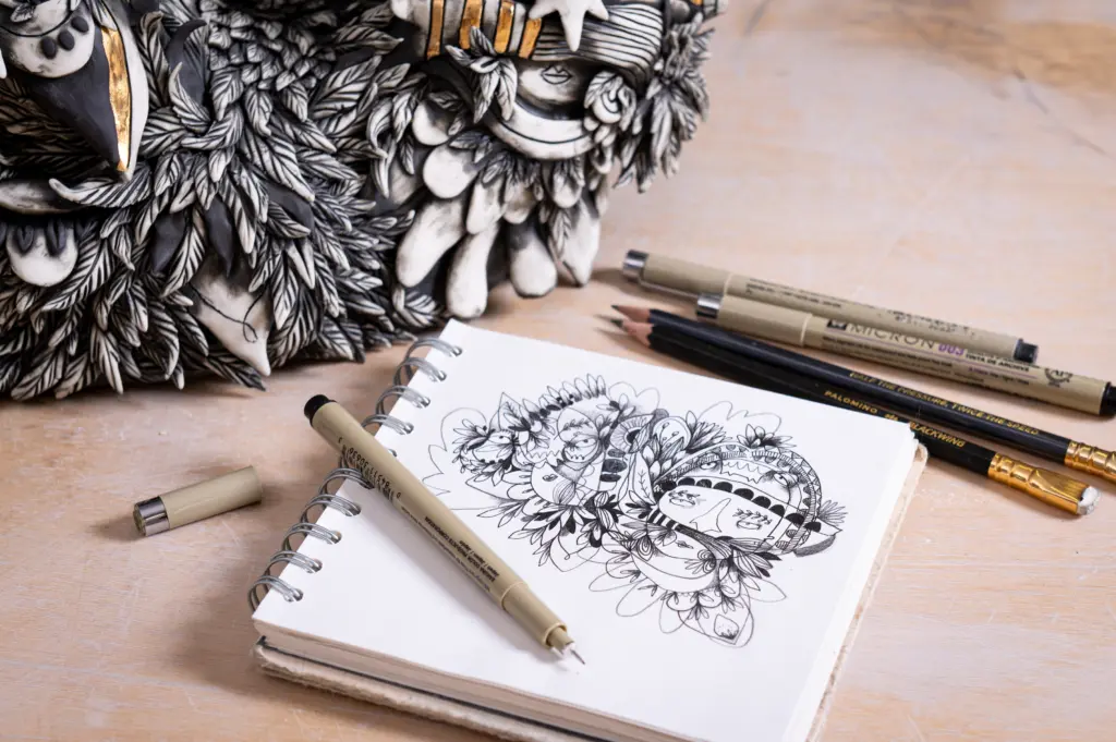 A photo of a sketchbook lying next to a ceramic sculpture. The sketchbook has an illustrative drawing of characters in a unique style and the sculpture is an interpretation of the drawing. There are pens lying on the table next to the sculpture and sketchbook.