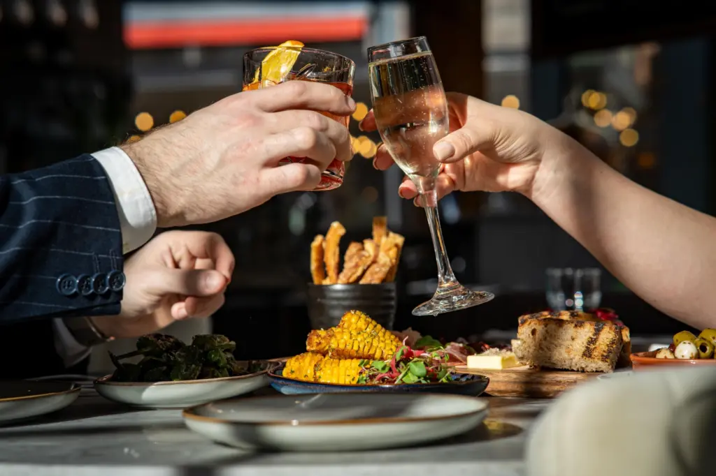Photo of two hands holding wine glasses and clinking their glasses together. They are at a table with steak and corn on plates. It all looks very classy.