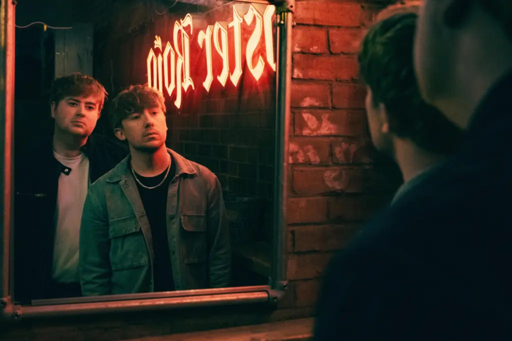 Two men looking in a mirror against a brick wall with a red neon light shining on their faces. The photo is grainy and moody.