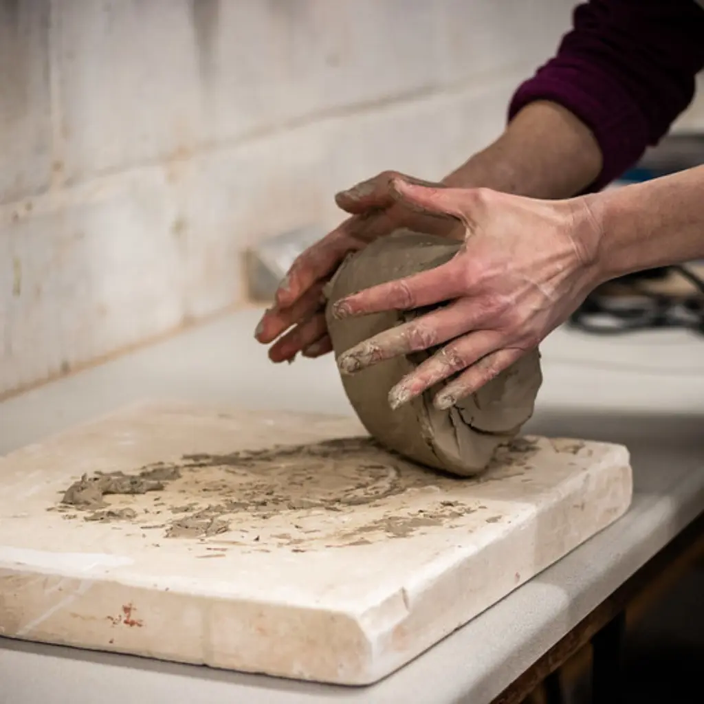 A student is kneading clay. Their hands are messy from the clay and they are mid-action.