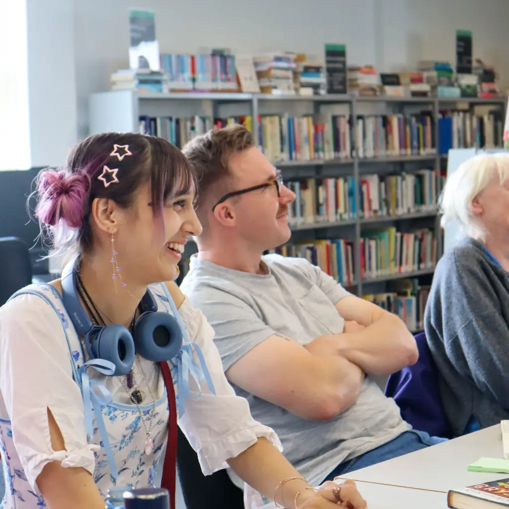 A girl is looking off-camera to the right, smiling. She is sat at a desk with writing books in front of her. She has pink hair styled in space buns. Next to her, a man with glasses is sitting back in his chair and also looking off-camera. They are both sat in front of bookshelves, in a library.