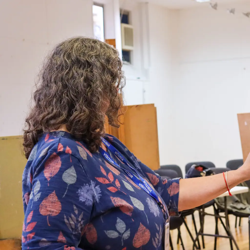 A lady with dark grey curly hair is stood at an easel drawing. She wears a patterned top, and is looking off camera, so you can't see her face.