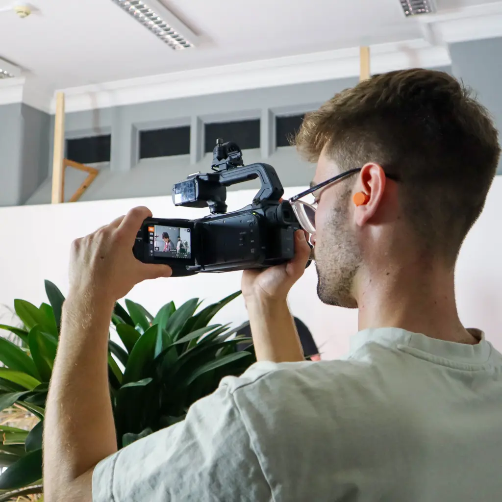 A young man with a video camera, filming something off-screen. He is turned away from us, concentrating on the camera screen. The room is light and bright, with plants dotted around.