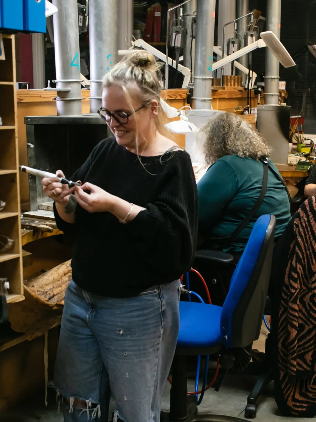 A woman with glasses and long blonde hair tied up in a bun stands in a small metals workshop, smiling whilst working with a small piece of silver.