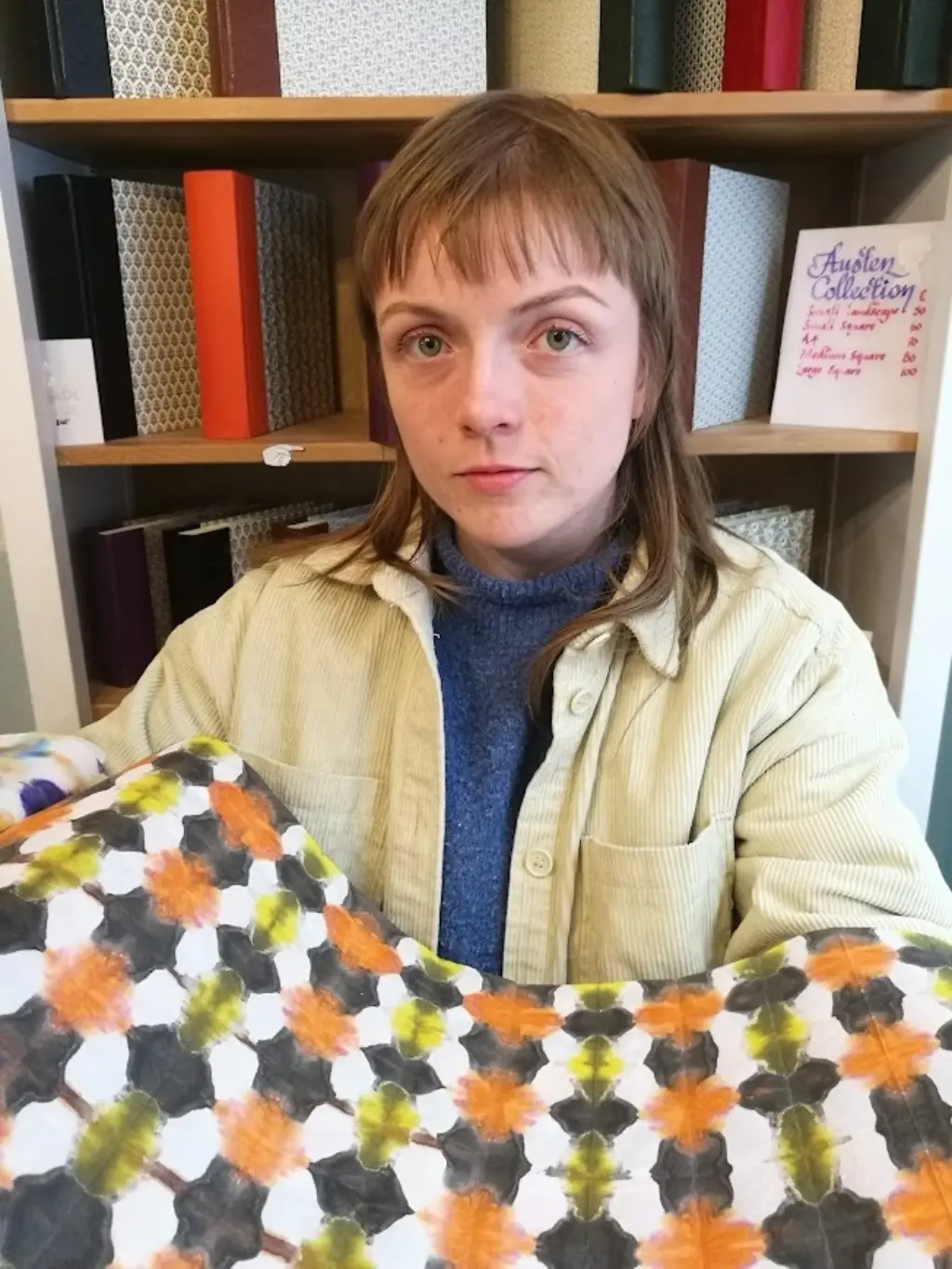 A portrait of Aoife Higgins. She is in a bookbinding studio, and is holding some colourful patterned paper.