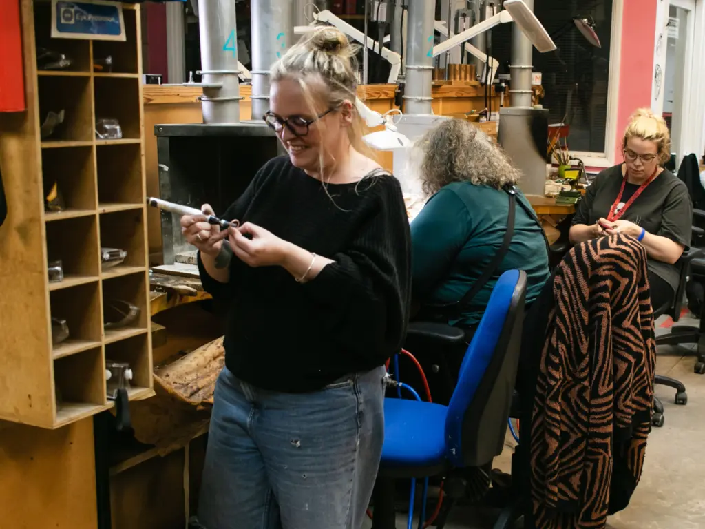 A woman with glasses and long blonde hair tied up in a bun stands in a small metals workshop, smiling whilst working with a small piece of silver.