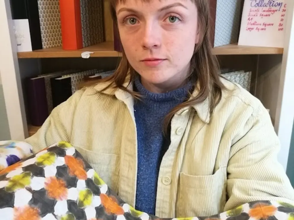 A portrait of Aoife Higgins. She is in a bookbinding studio, and is holding some colourful patterned paper.