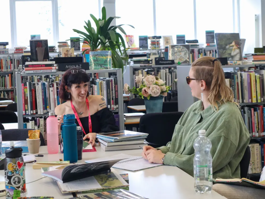 Two women sat at a desk in a library, surrounded by bookshelves and plants. In front of them are stacks of books and open notebooks that they are writing in. They are talking to one another and smiling.