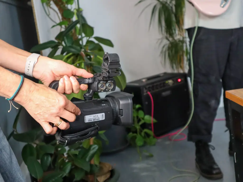 A person off-screen holds a video camera, filming a band in a photography studio.