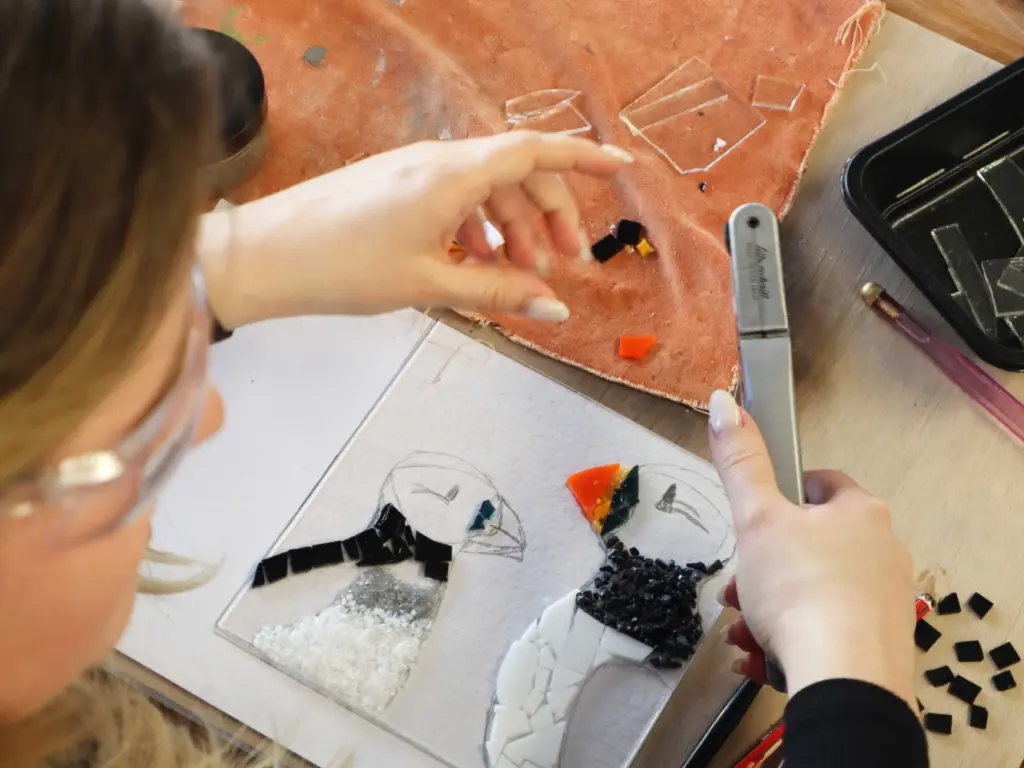 An over the shoulder shot of a woman working on a glass fused artwork of two puffins. She is cutting the next piece of glass to go on.