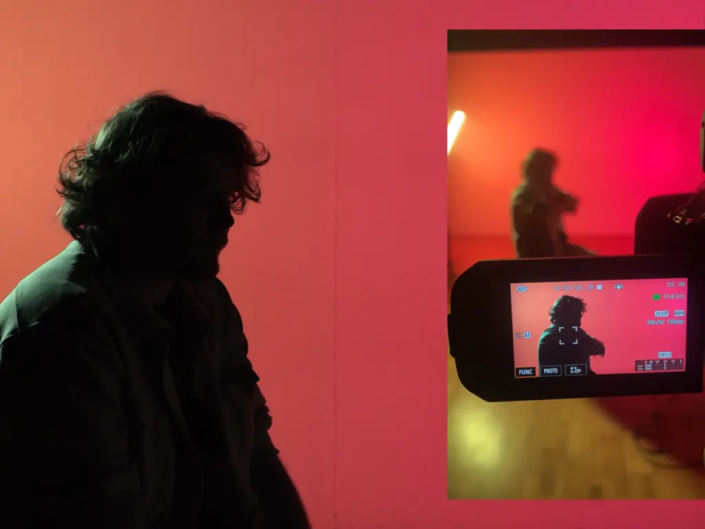 The silhouette of a man against a red background. The screen of a camera is in the foreground, filming the man.