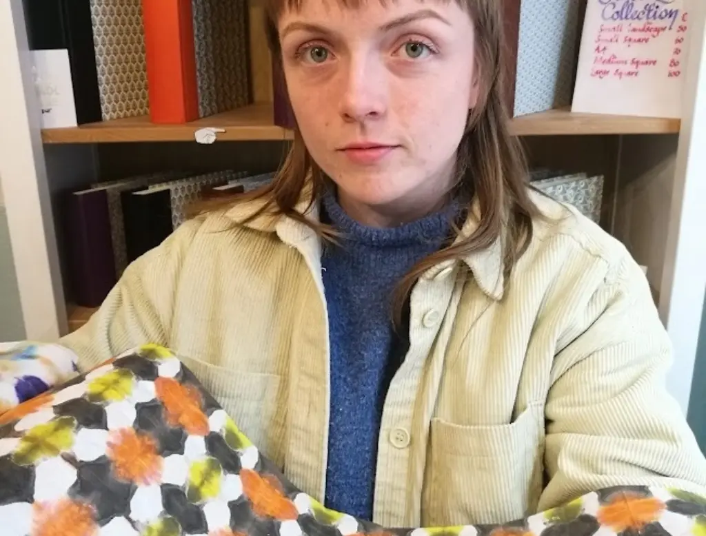 A portrait of Aoife Higgins. She is in a bookbinding studio, and is holding some colourful patterned paper.
