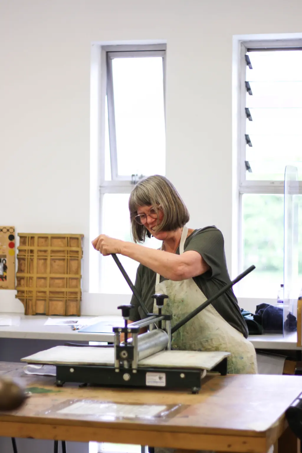 A lady with short grey hair using a printing press in a light and airy print workshop