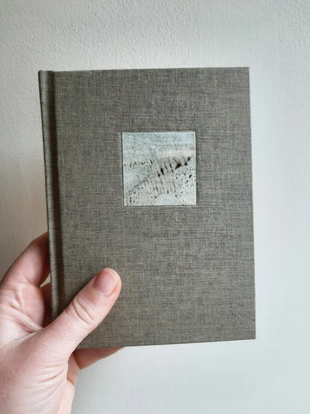 A hand is holding a small grey, cloth-bound book with a small picture on the front of it.