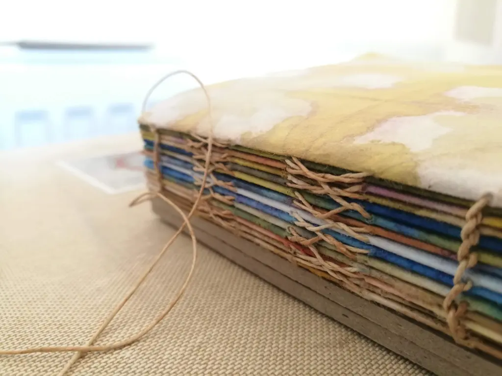 A close-up of a hand-stitched bound book, with decorative stitching on the side. The papers are different colours and look marbled.