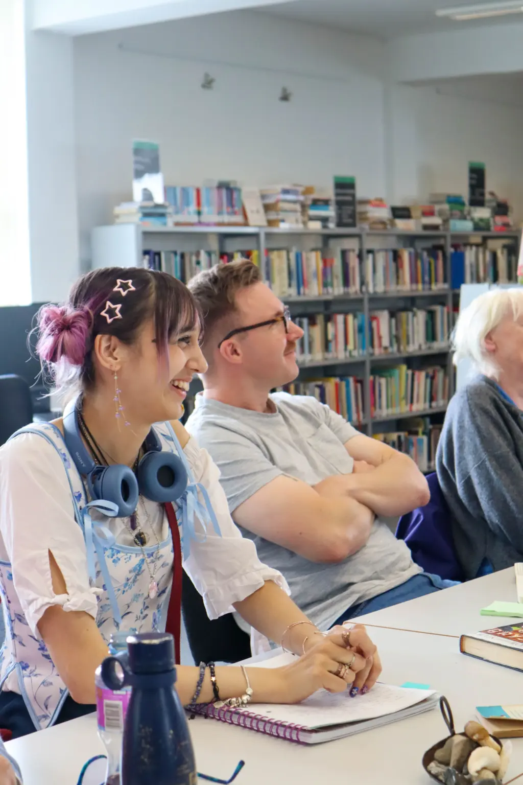 A girl is looking off-camera to the right, smiling. She is sat at a desk with writing books in front of her. She has pink hair styled in space buns. Next to her, a man with glasses is sitting back in his chair and also looking off-camera. They are both sat in front of bookshelves, in a library.