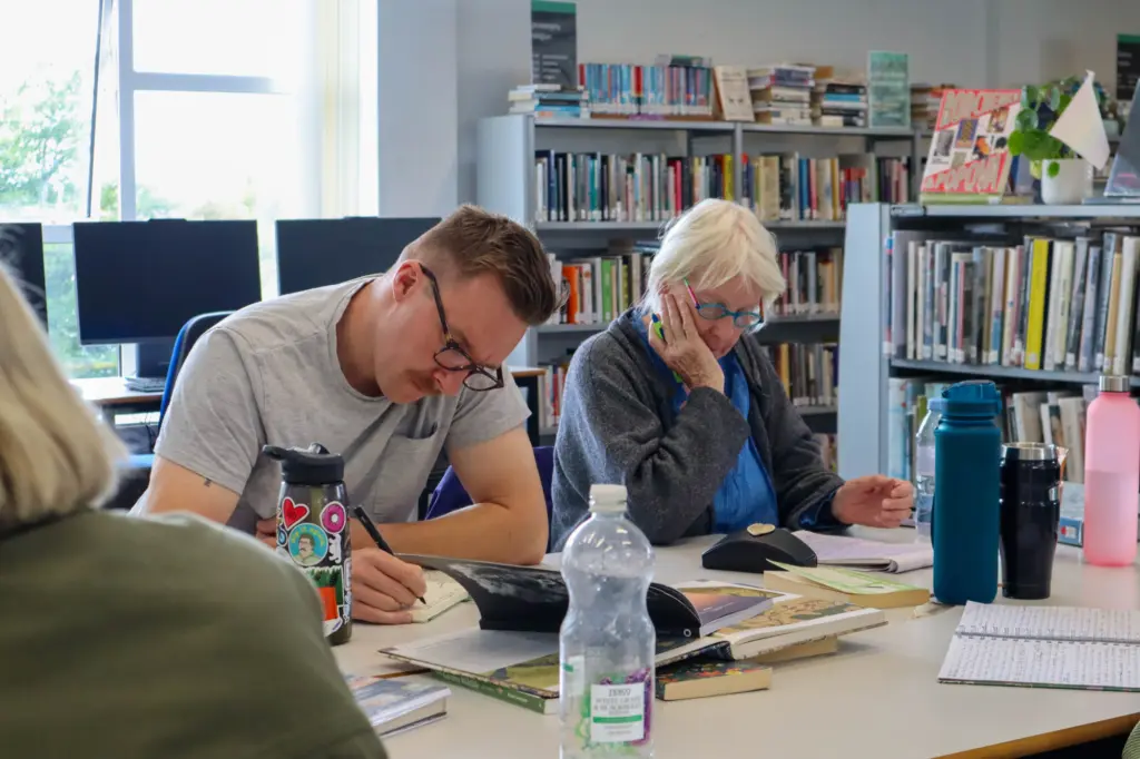 A man and a woman both sat at a desk, concentrating on books in front of them. They are writing in the books on the desk, and the lady is resting her head in her hand.