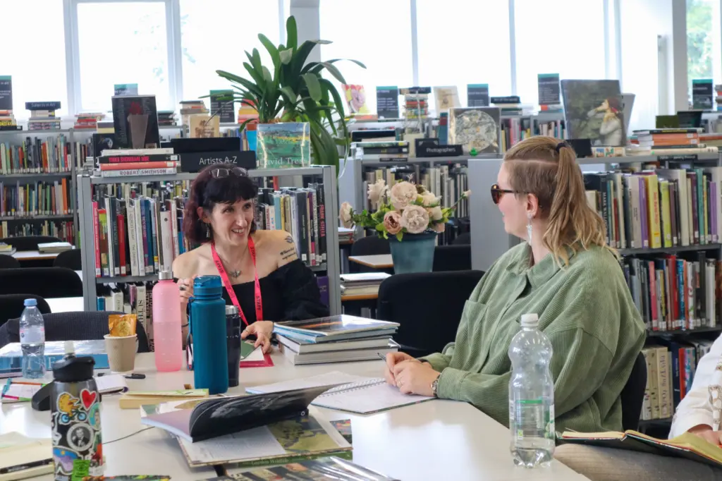 Two women sat at a desk in a library, surrounded by bookshelves and plants. In front of them are stacks of books and open notebooks that they are writing in. They are talking to one another and smiling.