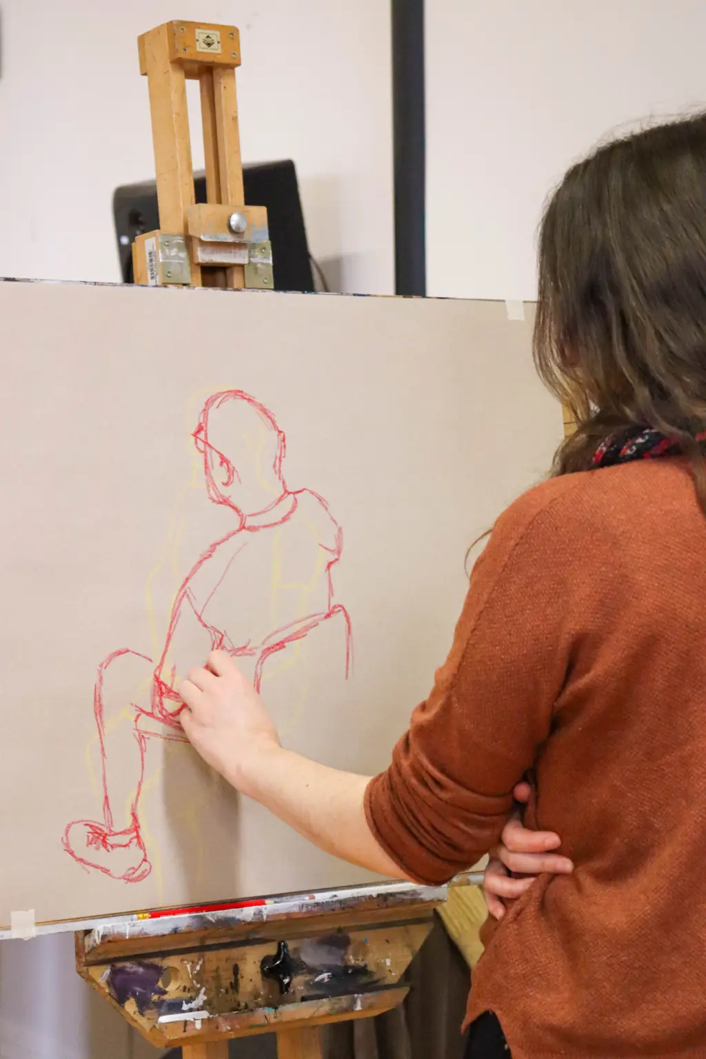 Looking over the shoulder of a woman in an orange jumper, you can see her drawing on a board at an easel. The drawing is a simple but accurate line drawing of a man sat in a chair.