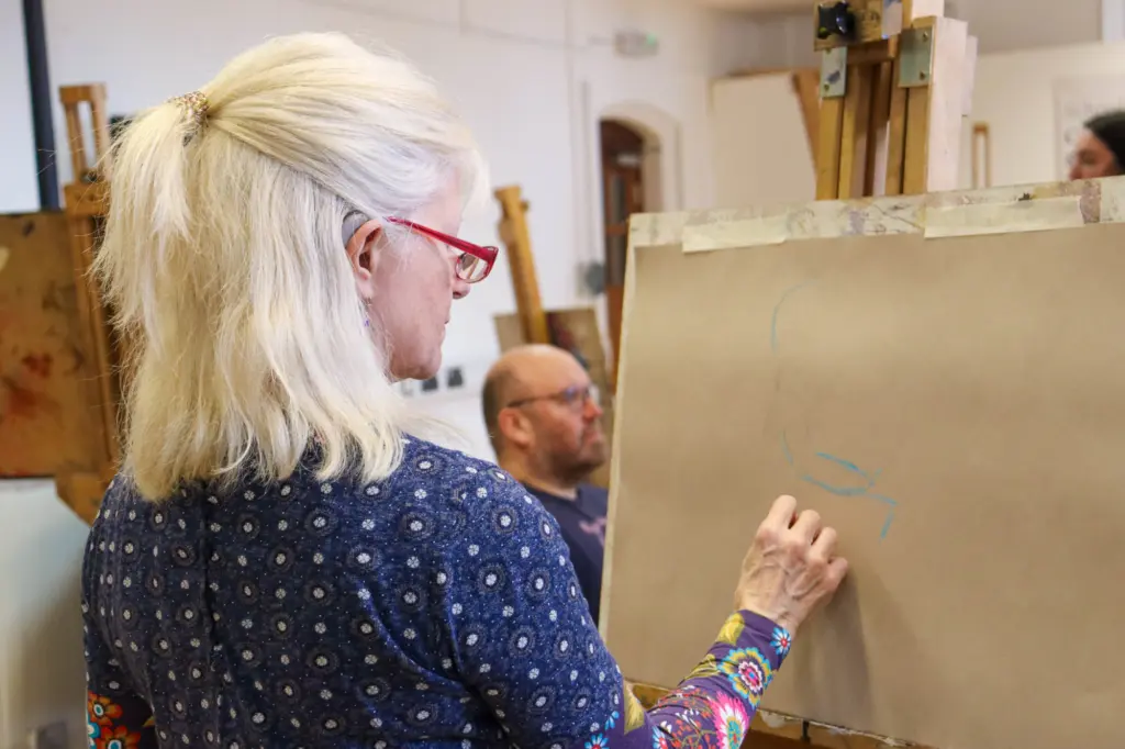 A lady with white hair and bright red glasses is focussed on drawing on a board in front of her. In the background, you can see the model sat down.