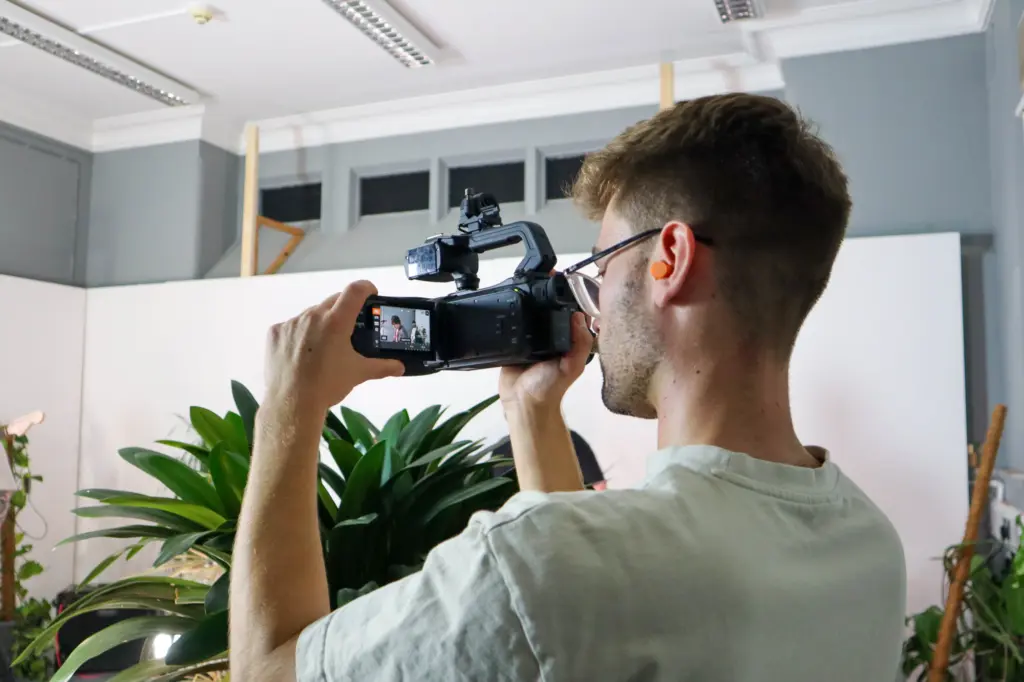 A young man with a video camera, filming something off-screen. He is turned away from us, concentrating on the camera screen. The room is light and bright, with plants dotted around.