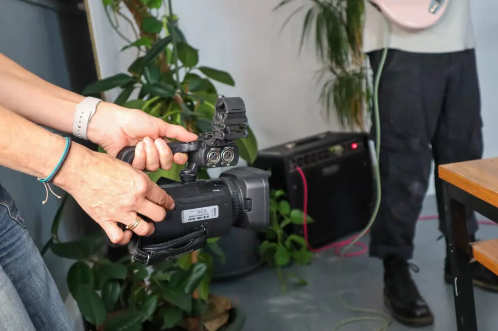 A person off-screen holds a video camera, filming a band in a photography studio.