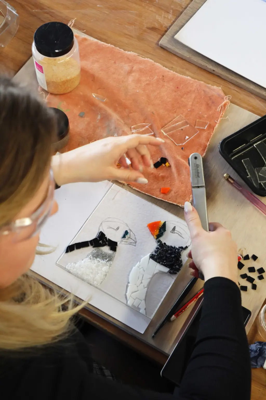 An over the shoulder shot of a woman working on a glass fused artwork of two puffins. She is cutting the next piece of glass to go on.