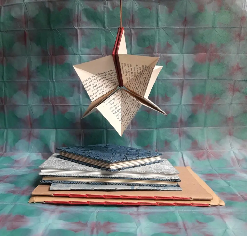 A pile of hand-made notebooks stacked up with a star-shaped, folded out book decoration hanging from above.