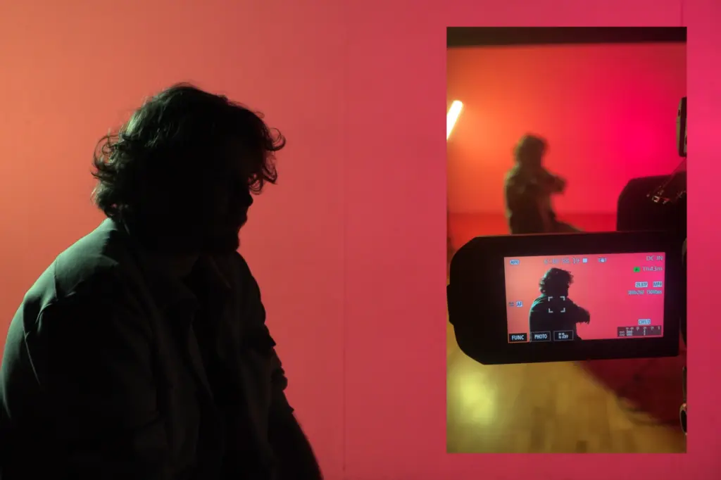 The silhouette of a man against a red background. The screen of a camera is in the foreground, filming the man.