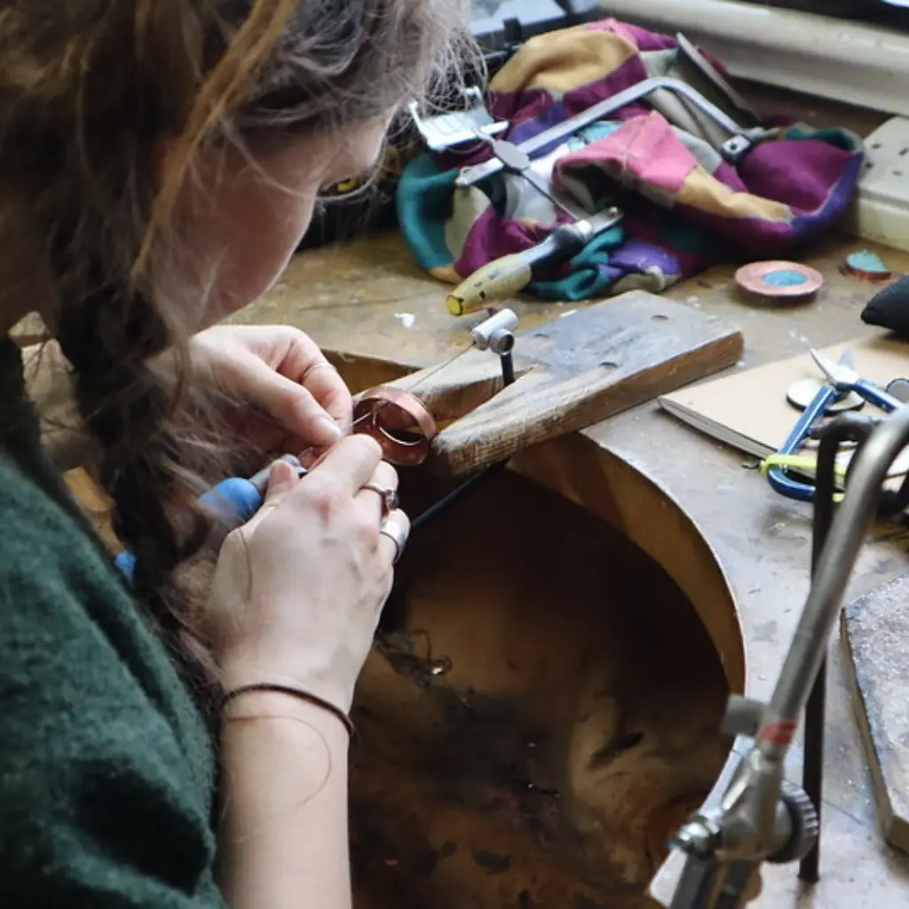 An over-the-shoulder shot of a young woman working at a small metals bench.