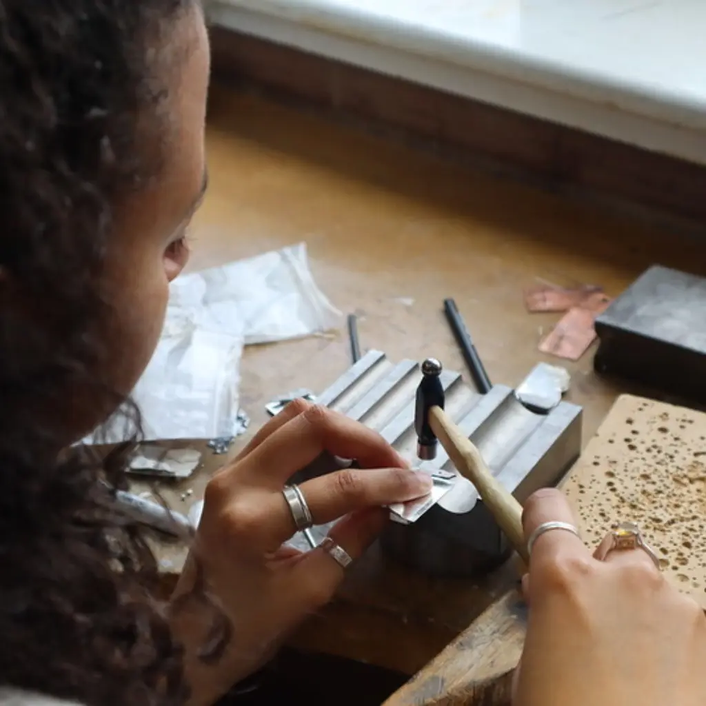 A young woman working at the small metals bench. She has silver rings on her fingers and is hammering another silver ring.