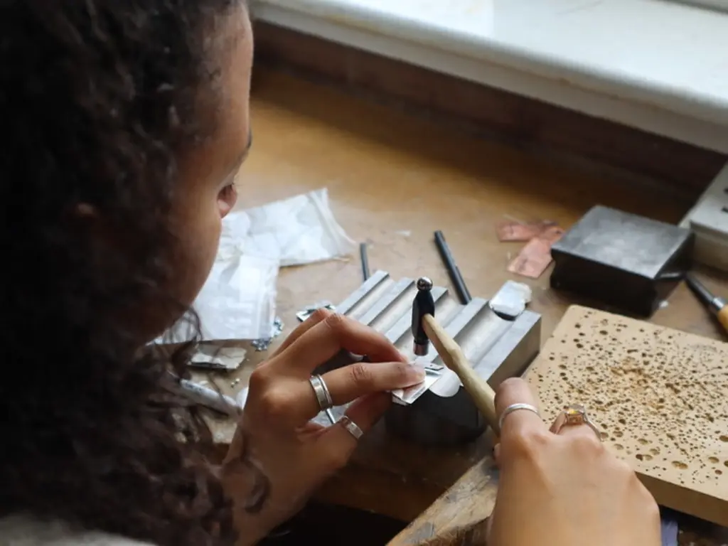A young woman working at the small metals bench. She has silver rings on her fingers and is hammering another silver ring.