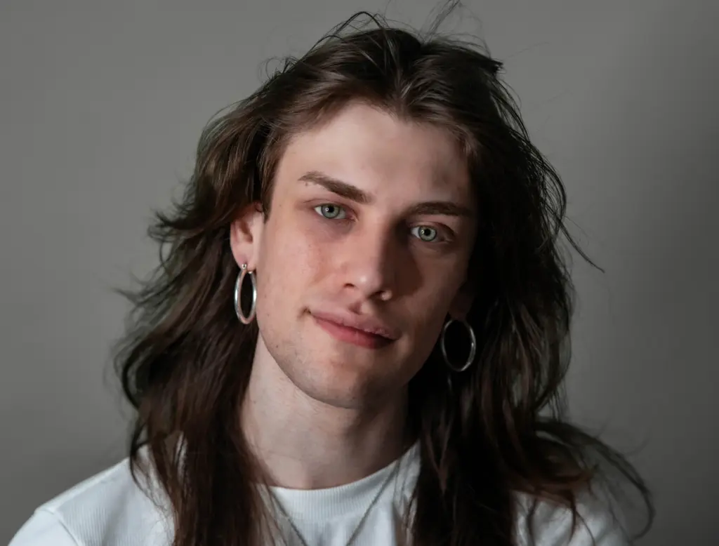A studio photo of a person with long brown hair and hoop earrings against a grey background.