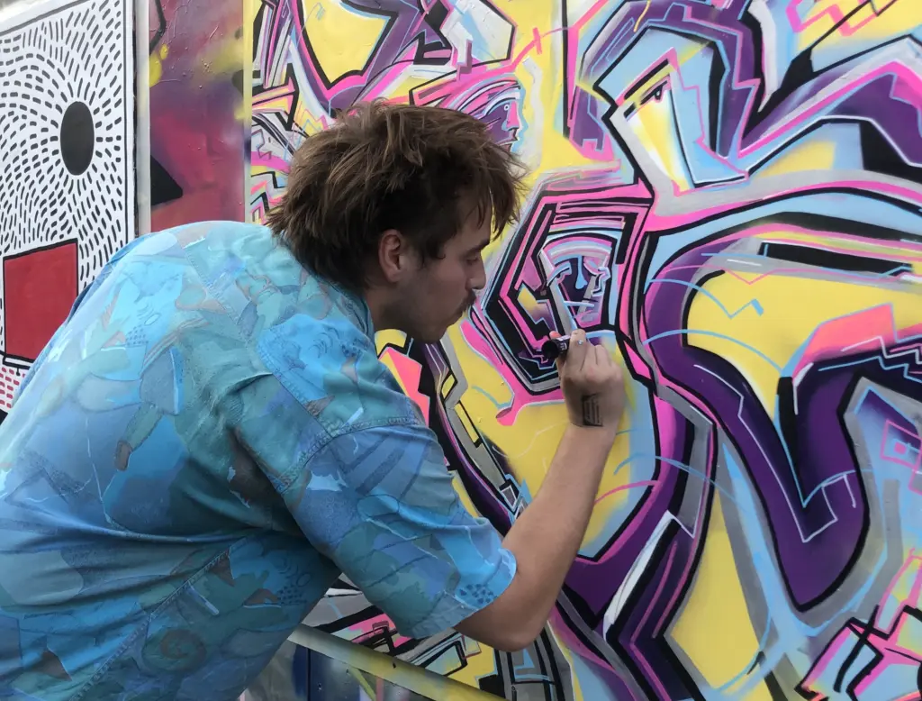 A man with a blue shirt and shaggy brown hair working on a street art-style mural.