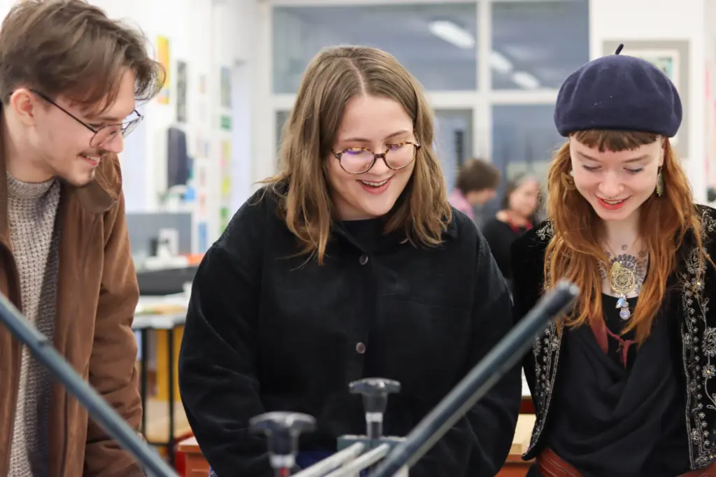 3 people all gathered around a table looking at work together and smiling.