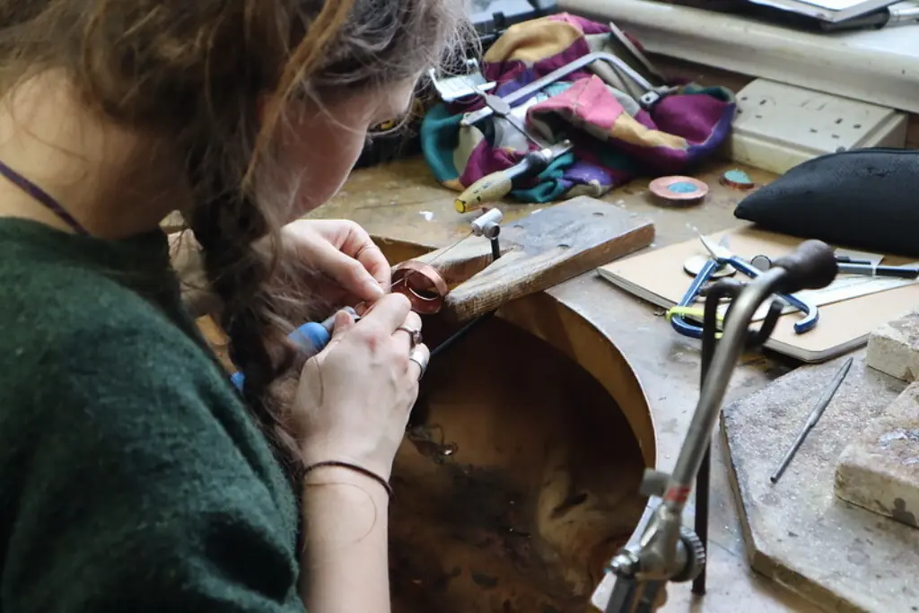 An over-the-shoulder shot of a young woman working at a small metals bench.